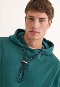 Sudadera azul verdosa con cierre de cordón que presenta un cordón de patrón en blanco y negro. Confeccionada con una tela suave y texturizada.
