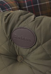 Olive green quilted fabric with a round brown leather label embossed with "Barbour." Features prominent stitching and a central button detail.