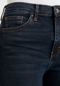 Vaqueros de mezclilla azul oscuro con un diseño clásico, cinco bolsillos y costuras naranjas en contraste a lo largo de las costuras y los bordes de los bolsillos.