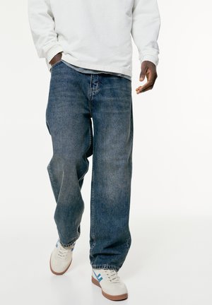 Baggy blue denim jeans with a faded wash, coupled with white sneakers featuring blue accents and beige soles, set against a plain background.
