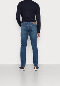 Niebieskie jeansy z denimu o prostym kroju, pięcioma kieszeniami i brązowym skórzanym akcentem na pasku. Połączone z ciemnoniebieską bluzką z długim rękawem i sportowymi butami.