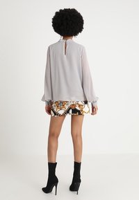 Lysegrå bluse med fallende ærmer, nøglehul detalje på ryggen, parret med mønstrede shorts med guld- og sorte accenter samt høje sorte støvler.