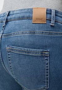 Close-up af blå denim jeans baglomme og linning med et syet tan lædermærke med teksten ONLY.