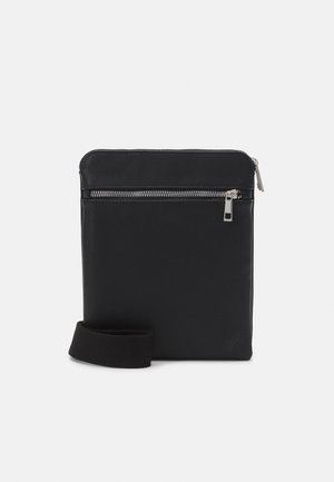 Sac bandoulière noir avec poche zippée à l’avant, détails de fermeture éclair argentés et bandoulière en tissu amovible sur fond blanc.