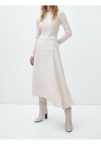 Femme portant un haut côtelé crème à manches longues, une jupe beige asymétrique et des bottes à talons hautes beige assorties sur un fond uni.