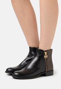 Bottes de cheville en cuir noir avec un dos en monogramme marron et des détails en zip doré, présentant une semelle plate et un design à bout arrondi.