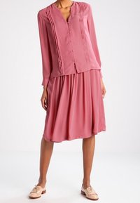 Woman wearing a long-sleeve pink blouse with pleats and matching knee-length skirt, paired with beige tassel loafers, standing against white background.