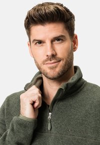 Pulover din fleece verde închis, cu gât cu fermoar, suprafață texturată și căptușeală interioară contrastantă. Prezintă un logo pe piept.