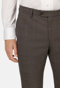 Pantaloni a quadri grigi con una texture liscia, dotati di chiusura a bottone e passanti per cintura, mostrati accanto a un polsino di una camicia bianca.
