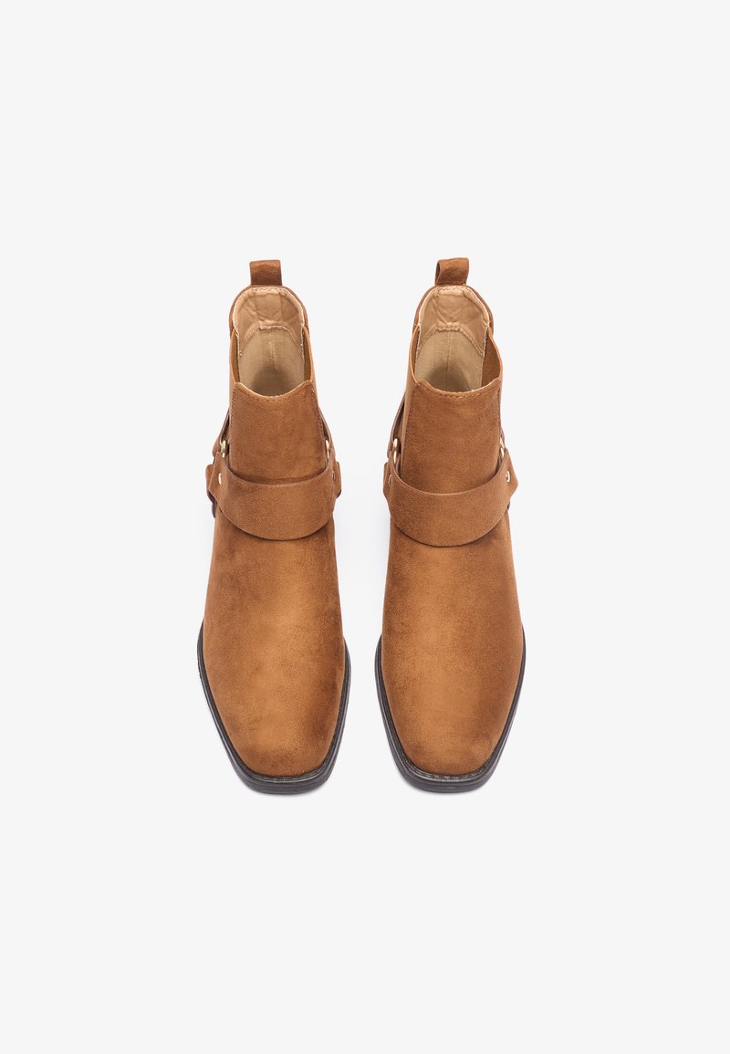 Botas de tobillo de gamuza marrón con punta cuadrada, detalle de correa lateral y tiradores en el empeine para facilitar su uso. Textura suave.