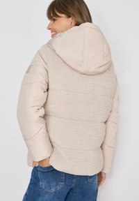 Veste matelassée beige clair avec un dos et une capuche en peluche, panneaux cousus et manches lisses. Présente un patch logo sur l'épaule.