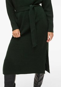 Kobieta w ciemnozielonej dzianinowej sukience z paskiem w talii i długimi rękawami, zestawiona z czarnymi wysokimi butami za kolano.