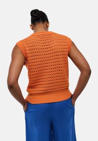 Pull sans manches en tricot orange avec un motif ajouré, col et ourlet côtelés, associé à un pantalon bleu. Détail visible au dos.