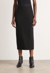 Jupe midi noire en tissu extensible avec une texture lisse, dotée d'une large ceinture. Associée à des bottines noires à bout pointu.