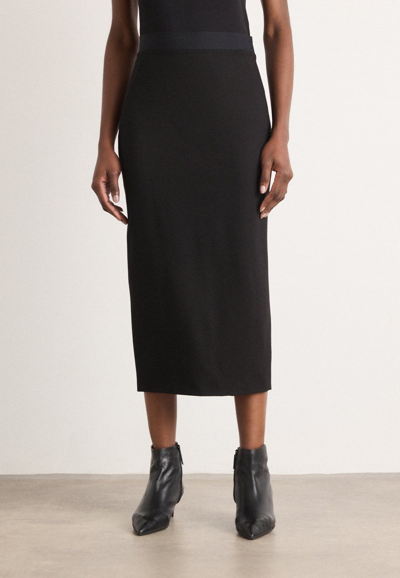 Jupe midi noire en tissu extensible avec une texture lisse, dotée d'une large ceinture. Associée à des bottines noires à bout pointu.