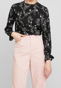 Svart blus med blommönster i nyanser av blått, rosa och grönt; har hög hals och volangärmar, matchad med ljusrosa byxor.