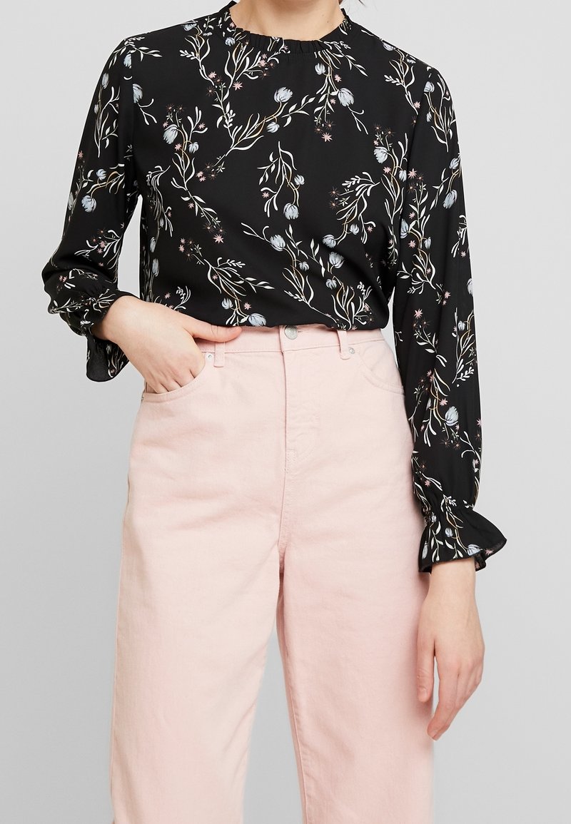 Svart blus med blommönster i nyanser av blått, rosa och grönt; har hög hals och volangärmar, matchad med ljusrosa byxor.