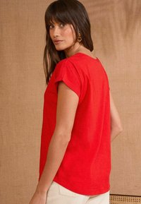 Chemise rouge à manches courtes avec un col rond et une coupe décontractée. Elle présente une texture lisse et un ourlet arrondi, portée avec un pantalon de couleur claire.