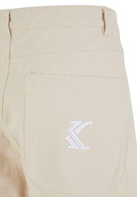 Pantaloni din bumbac bej, cu un buzunar posterior dotat cu un logo alb brodat. Materialul are o textură care subliniază durabilitatea.