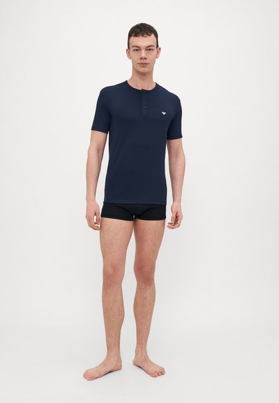 Chemise à manches courtes bleu marine avec une patte de boutonnage, texture côtelée et petit logo sur la poitrine gauche, portée avec des slips noirs.