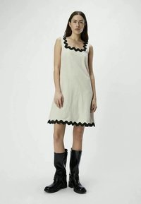 Woman wearing a sleeveless cream dress with black wavy trim and black knee-high boots, standing against a plain light background.
