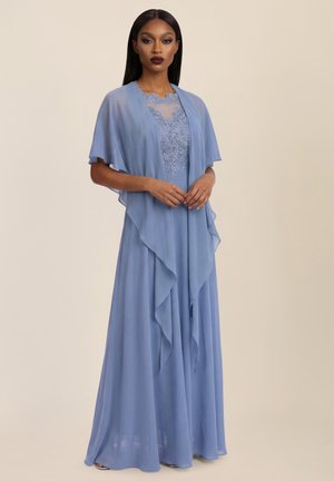 Frau steht mit verschränkten Händen und trägt ein fließendes, staubblaues Kleid mit Spitzenoberteil und passendem, transparentem, drapiertem Schal.