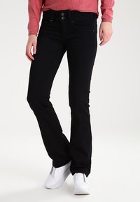 Pantalones vaqueros negros de corte acampanado hechos de denim, con un cierre de dos botones y bolsillos delanteros, combinados con zapatillas blancas y una camiseta de manga larga rosa.