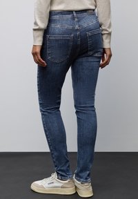 Jean skinny en denim bleu foncé, avec taille mi-haute, deux poches arrière et décoloration subtile. Assortis à des baskets blanches.