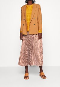 Beige Doppelreißverschluss-Blazer über einem strukturierten gelben Strickoberteil, kombiniert mit einem gemusterten Maxirock in Orange und Weiß, abgerundet mit beigen Schiebesandalen.