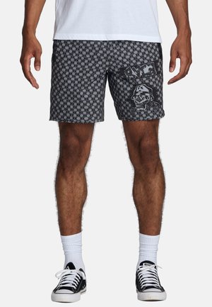Costumi da bagno neri a fantasia con grafica di teschio, con vita regolabile tramite cordino e tasche laterali. Indossati con sneakers nere e calzini bianchi.
