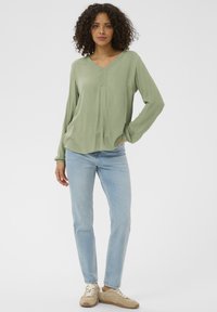 Blouse vert clair à manches longues avec un col en V et un détail de pli subtil, associée à un jean droit bleu clair et des baskets beiges.