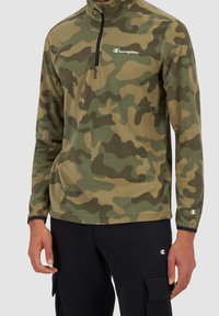 Tarnungs-Fleece-Pullover mit hohem Kragen, schwarzem Reißverschluss, langen Ärmeln und weißem gesticktem Logo auf der Brust. Strukturiert Oberfläche in Grün und Braun.