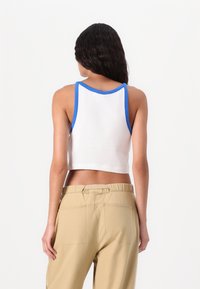 Hvid cropped tanktop med blå kant, der har en rund halsudskæring og brede stropper. Sammen med beige høj talje bukser. Glat stoftekstur.