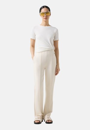 Kvinde iført hvid tætsiddende kortærmet top, cremefarvede bukser med høj talje og sømme, store solbriller med gultonede glas og cremefarvede sandaler.