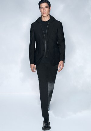Black tailored suit jacket with a hood and a front zipper, paired with a black top and trousers. Smooth texture, sleek silhouette, and sturdy shoes.