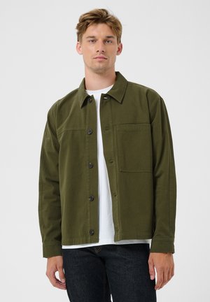 Joven con una chaqueta abotonada verde oliva sobre una camiseta blanca y vaqueros oscuros, de pie frente a un fondo claro y sencillo.