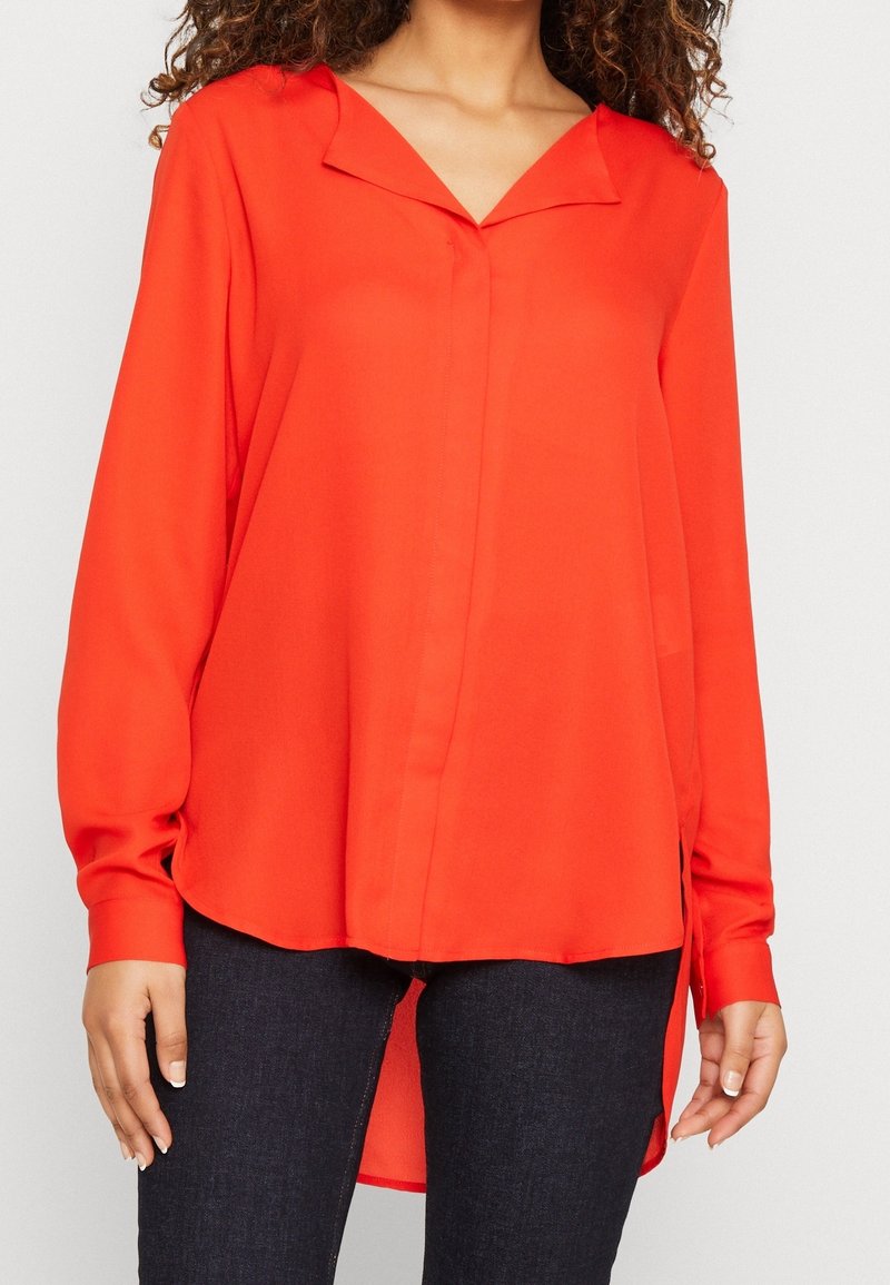 Blusa rossa a maniche lunghe con doppio colletto, patta frontale e spacchi laterali. Tessuto leggero con una superficie liscia, indossata con jeans scuri.