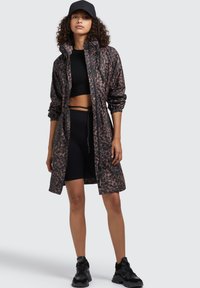 Giacca lunga e leggera con stampa leopardata marrone e nera, caratterizzata da una chiusura con zip, cappuccio regolabile con coulisse e polsini elasticizzati, abbinata a un top corto nero e pantaloni corti.