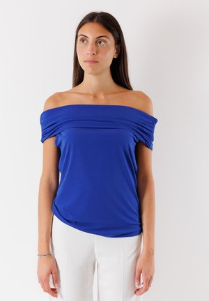 Top blu reale a spalle scoperte con dettagli arricciati, realizzato in tessuto morbido. Indossato con pantaloni bianchi, mostra un taglio rilassato.