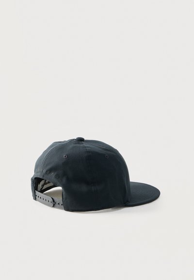 Μαύρο καπέλο snapback που ακουμπά σε επίπεδη επιφάνεια, φωτογραφημένο από την πίσω αριστερή πλευρά, σε απλό ανοιχτό φόντο.