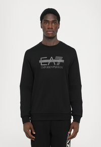 Svart hettegenser med en glanset grå "EA7 Emporio Armani" logo på brystet, laget av mykt, teksturert stoff med en løs passform.