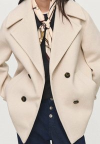Cappotto di lana beige doppiopetto con grandi bottoni neri e rever a pipistrello, abbinato a una sciarpa di seta con motivo crema e bordeaux.