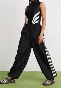 Bodysuit negro sin mangas con rayas curvas blancas, combinado con pantalones anchos negros con rayas verticales blancas y zapatos de slip-on con estampado animal.
