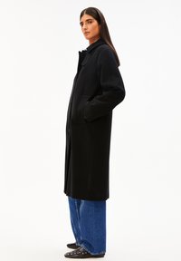 Frau im Profil stehend, trägt einen langen schwarzen Mantel, blaue weite Jeans und schwarze, mit Nieten besetzte Slipper vor einem weißen Hintergrund.