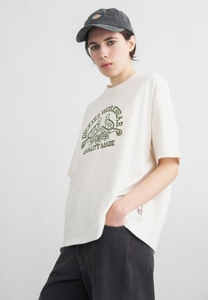 Noor inimene, kellel on seljas valge Dickiese graafilise mustriga T-särk, mustad püksid ja tumehall müts, poseerib ühe käega taskus.