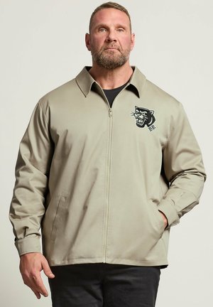 Mand iført beige lynlåstrøje med sort panterlogo på venstre bryst og sorte bukser, stående mod en lys baggrund.