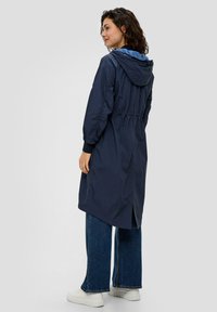 Granatowy długi płaszcz z kapturem, z wcięciem w talii i elastycznymi mankietami. Posiada rozcięty dół i wykonany jest z lekkiego materiału, zestawiony z szerokimi dżinsami.