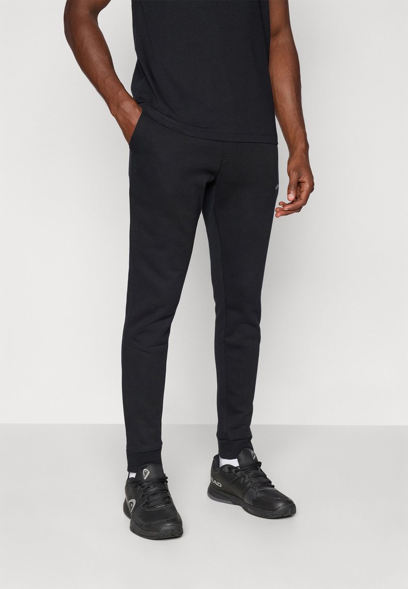 Pantalons de survêtement noirs fuselés avec des poignets élastiques, fabriqués à partir d'un tissu lisse. Associés à des chaussures de sport noires avec des accents gris.