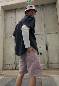 Grå teksturert kortermet skjorte over en hvit langermet skjorte, kombinert med lyse rosa shorts med logo på lommen; iført en rosa bøttehatt.
