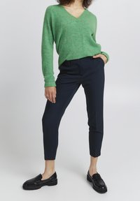 Zielony sweter w serek wykonany z miękkiego dzianiny, w połączeniu z ciemnonawigacyjnymi spodniami o dopasowanym kroju i czarnymi butami wsuwanymi.